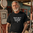 A man with a gray beard and tattoos stands in front of a vintage gas station pump wearing a black t-shirt with the white printed text 'Don't be a fucking fascist.'.