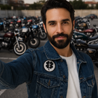 A man with a beard takes a selfie while wearing a blue denim jacket with a circular black and white four-leaf clover patch on the chest, standing in front of a row of parked motorcycles.