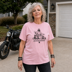 A woman wearing a light pink graphic t-shirt stands in a residential driveway with a black motorcycle parked behind her. The t-shirt features a black graphic of Jesus with a plow and the text 'This weary heart would be satisfied in happiness, peace and quiet from God alone'.