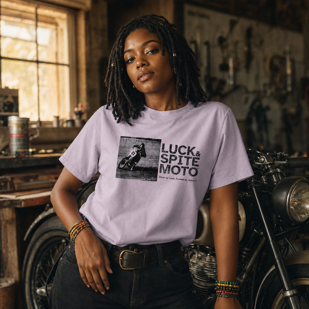A woman wearing a light lavender graphic t-shirt with a black and white 'Luck & Spite Moto' print, standing in a rustic motorcycle workshop.