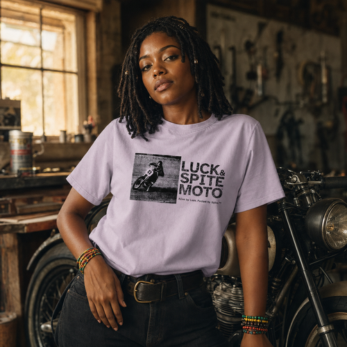 A woman wearing a light lavender graphic t-shirt with a black and white 'Luck & Spite Moto' print, standing in a rustic motorcycle workshop.